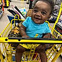 baby, shopping_cart, smiling, child, store, aisle, clothing, blue_shirt, yellow_shorts, happy, infant, curly_hair, retail, floor, product_shelves, yellow_cart, toys, portrait, indoor, cute
