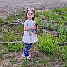 Isabella is registered to the contest to win money with this photo: bow, can, casual_clothing, child, curly_hair, daylight, dirt, exploration, forest_edge, girl, grass, greenery, leggings, nature, outdoor, plants, shirt, standing, tree, young_child