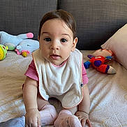 Maya participe au concours pour gagner de l'argent avec cette photo : baby, bed, bib, child, curious, cute, expression, face, furniture, home, indoor, person, pillow, pink, plush_toy, red, sitting, socks, soft_toys, white