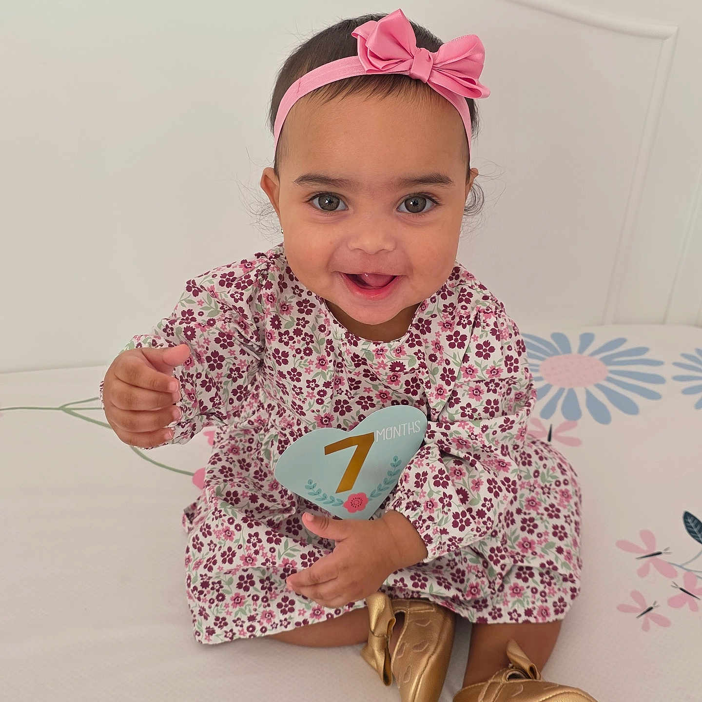 Alaia is registered to the contest to win money with this photo: baby, child, girl, smile, headband, pink_bow, floral_dress, golden_shoes, bed, happy, portrait, indoor, cute, infant, seven_months, sitting, person, young_child, playful, young_baby
