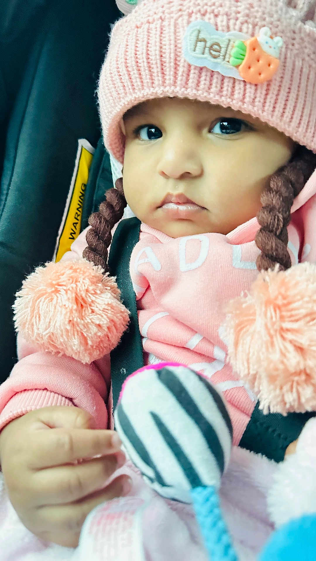 Laya joined the competition — help win amazing prizes! baby, child, pink_hat, pom_poms, car_seat, toy, striped_toy, hand, clothing, face, cute, infant, warm_clothing, braids, portrait, indoors, seatbelt, soft_toy, curious, close_up