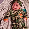 newborn, baby, infant, child, smiling, face, portrait, lying_down, blanket, striped_blanket, camouflage, outfit, hat, clothing, pacifier, bed, indoors, happy, close_up, photography