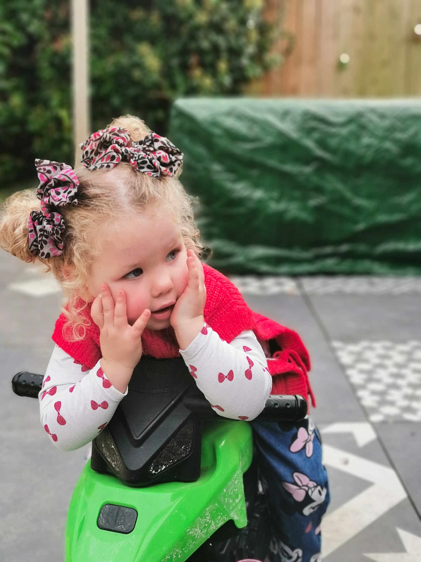 Azélïa participe au concours pour gagner de l'argent avec cette photo : child, girl, curly_hair, hair_bows, toy_bike, green_toy, outdoor, garden, red_clothing, long_sleeve_shirt, patterned_pants, playful, thoughtful, blonde, young_child, hands_on_face, cute, daylight, pavement, casual