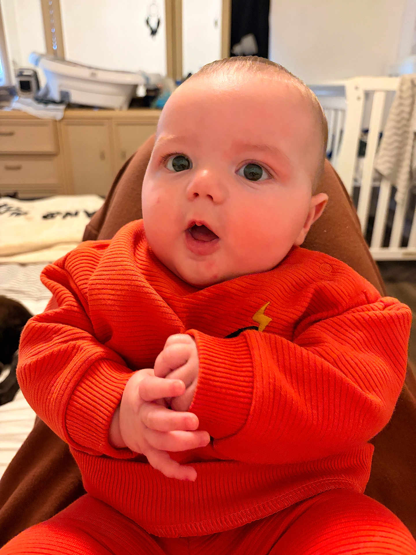 Colton joined the competition — help win amazing prizes! baby, infant, child, portrait, close_up, orange_outfit, sweater, hands, big_eyes, surprised_expression, crib, bedroom, dresser, blanket, indoor, soft_lighting, cute, skin, sitting, person