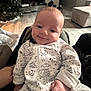Colton is registered to the contest to win money with this photo: baby, infant, smiling, onesie, pajamas, cozy, living_room, christmas_tree, tv, couch, wood_floor, blanket, hand, face, eyes, cheeks, indoor, parent_hand, portrait, relaxed