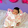 baby, infant, onesie, pink_hearts, stuffed_animal, bedspread, floral_pattern, purple_wall, balloon_letters, pink_balloon, soft_toy, sleepy, cute, newborn, indoors, cozy, relaxed, portrait, child, small