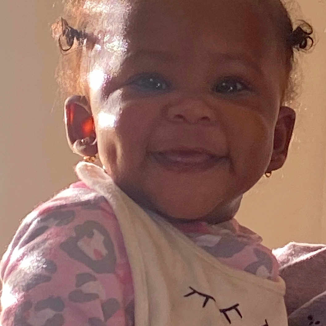 Makiyha is registered to the contest to win money with this photo: apparel, baby, bib, child, cute, dimples, earrings, expression, face, hair_buns, happy, indoor, light, onesie, person, portrait, shadow, skin, smile, toddler