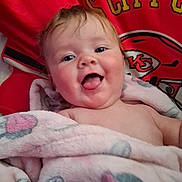 Athena is registered to the contest to win money with this photo: baby, child, blanket, pink_blanket, tongue_out, happy, smiling, face, chubby_cheeks, red_background, sports_logo, kc_chiefs, infant, portrait, cute, wrapped, indoors, young_child, soft_texture, closeup