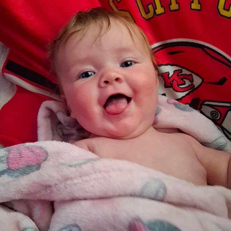 Athena is registered to the contest to win money with this photo: baby, child, blanket, pink_blanket, tongue_out, happy, smiling, face, chubby_cheeks, red_background, sports_logo, kc_chiefs, infant, portrait, cute, wrapped, indoors, young_child, soft_texture, closeup