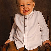Camille participe au concours pour gagner de l'argent avec cette photo : toddler, child, smile, white_shirt, brown_pants, wooden_chair, indoors, portrait, happy, seated, young_child, cute, face, hands, buttoned_shirt, warm_lighting, brown_background, long_sleeves, casual_clothing, small_child