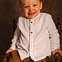 toddler, child, smile, white_shirt, brown_pants, wooden_chair, indoors, portrait, happy, seated, young_child, cute, face, hands, buttoned_shirt, warm_lighting, brown_background, long_sleeves, casual_clothing, small_child