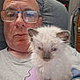 man, glasses, bald, kitten, cat, fluffy, indoor, chair, sitting, shelf, figurines, vacuum_cleaner, gray_shirt, closeup, pet, home, portrait, furniture, relaxed, casual