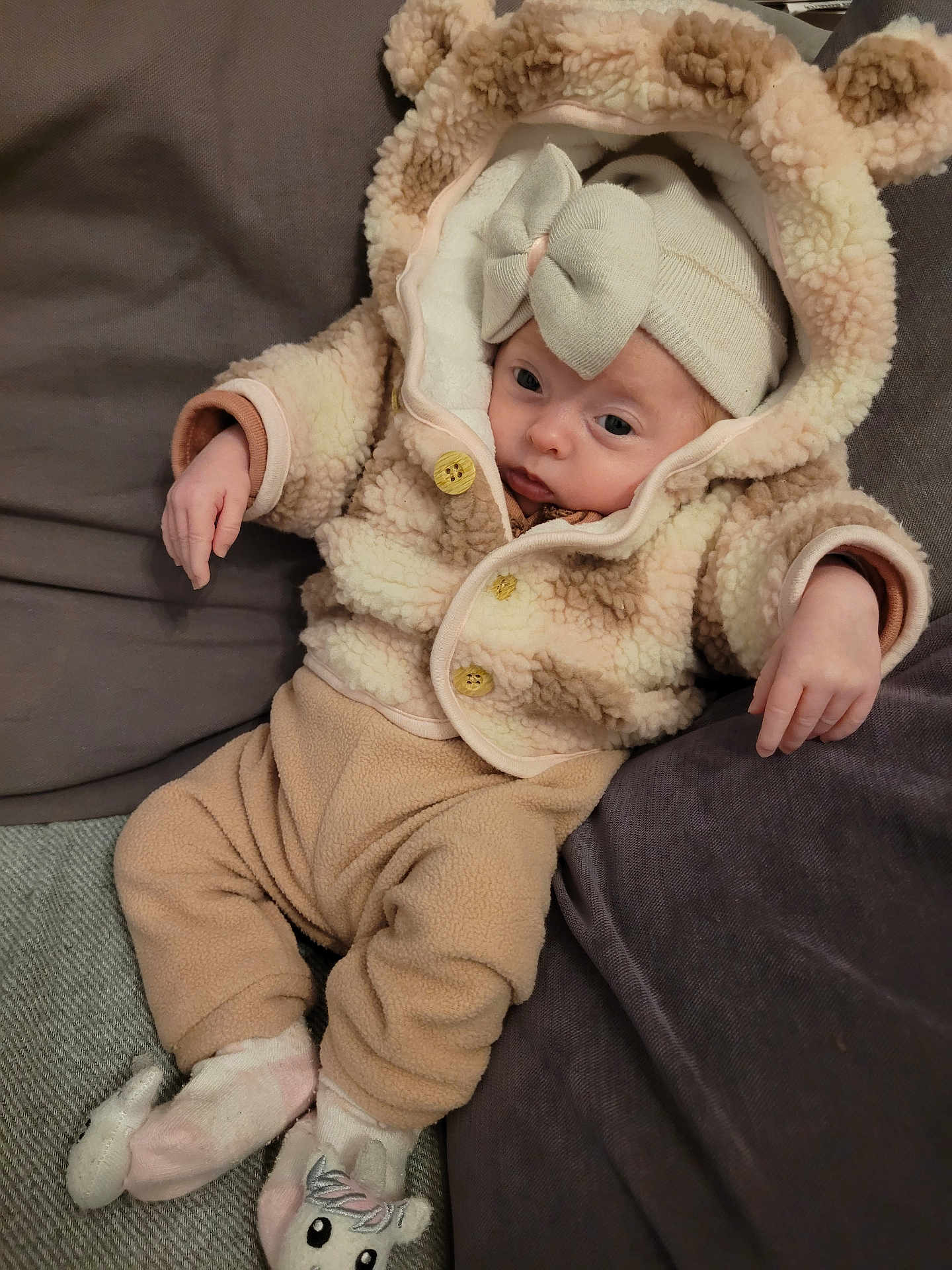 Sofia participe au concours pour gagner de l'argent avec cette photo : baby, child, infant, hooded_jacket, bear_ears, bow_hat, unicorn_slippers, couch, cushion, cozy_clothing, soft_fabric, relaxed, indoors, person, cute, warm, sleepwear, pink, beige, comfortable
