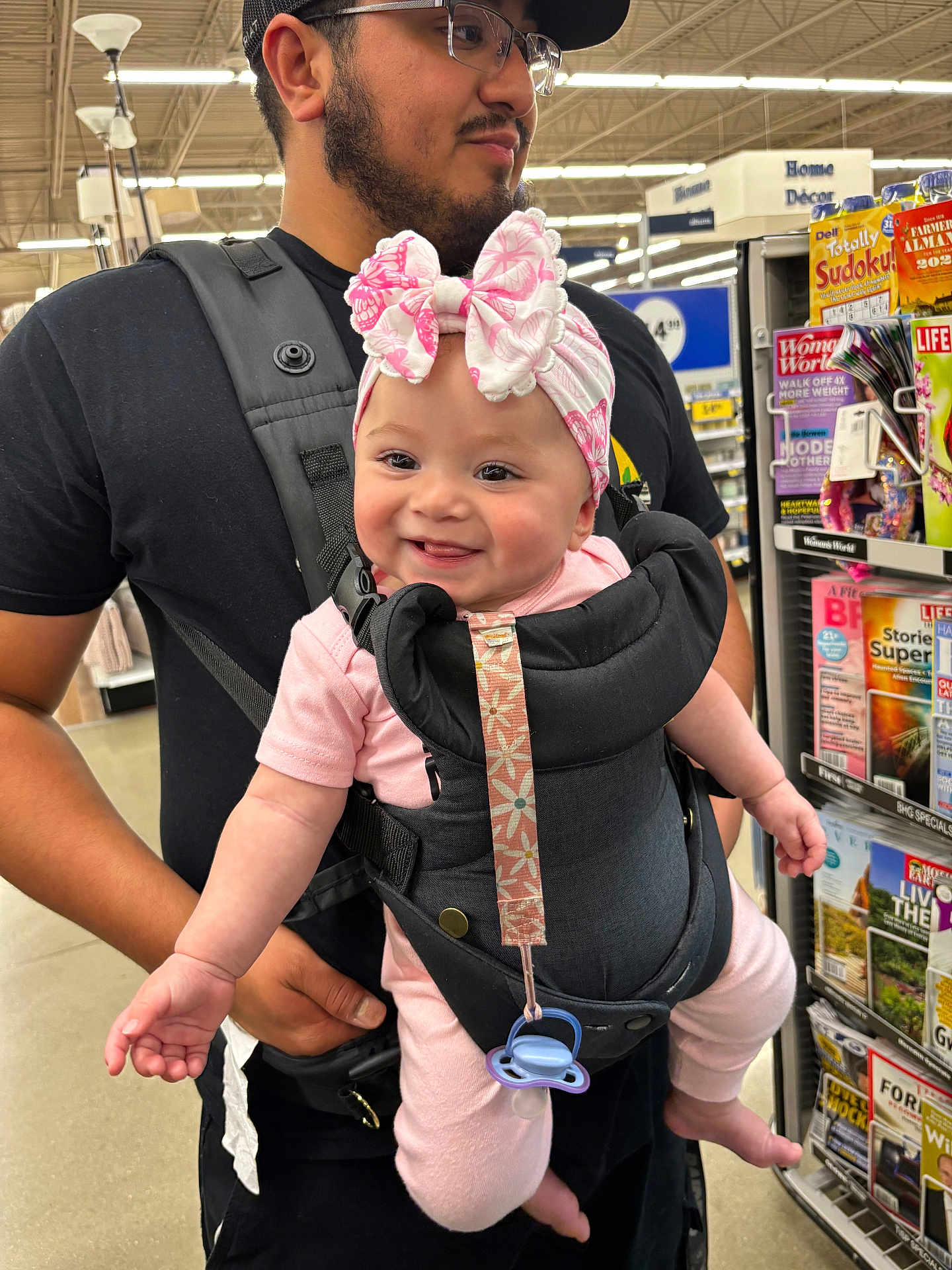 Ezme is registered to the contest to win money with this photo: baby, man, baby_carrier, pink_clothing, headband, smiling, indoor, store, magazines, shelf, shopping, adult, black_shirt, face, person, retail, holding, pacifier, happy, beard