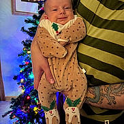 Angus is registered to the contest to win money with this photo: baby, infant, person, hand, tattoo, striped_shirt, christmas_tree, holiday, lights, decorations, onesie, polka_dot, holly, smile, indoor, holding, festive, warm_lighting, portrait, celebration