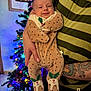baby, infant, person, hand, tattoo, striped_shirt, christmas_tree, holiday, lights, decorations, onesie, polka_dot, holly, smile, indoor, holding, festive, warm_lighting, portrait, celebration