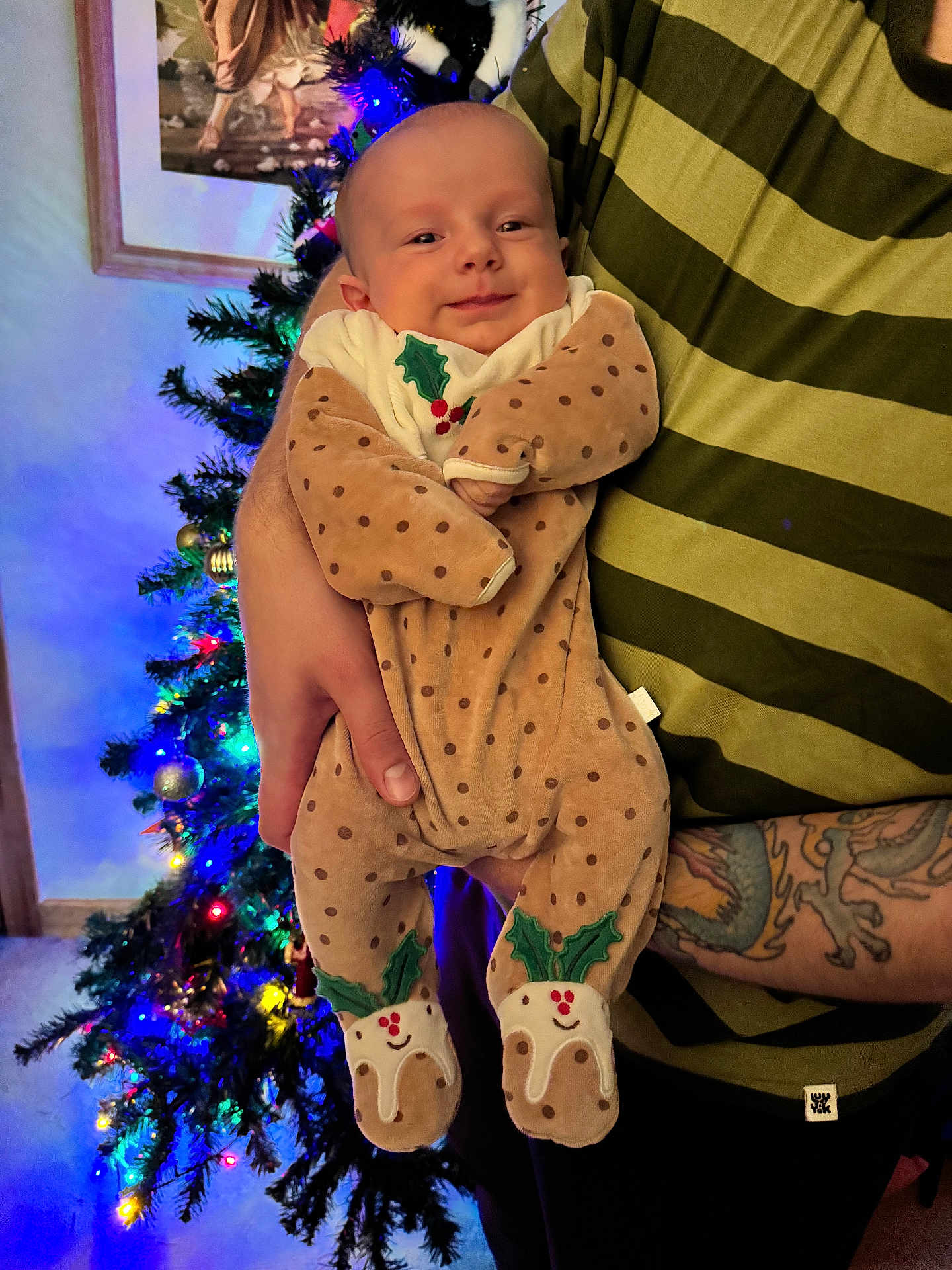 Angus is registered to the contest to win money with this photo: baby, infant, person, hand, tattoo, striped_shirt, christmas_tree, holiday, lights, decorations, onesie, polka_dot, holly, smile, indoor, holding, festive, warm_lighting, portrait, celebration