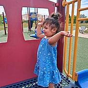 Rory joined the competition — help win amazing prizes! toddler, child, playground, blue_dress, barefoot, outdoor, play_equipment, happy, smiling, girl, daylight, summer, fun, young_child, plastic_structure, yellow_railings, red_wall, grass, bench, sky
