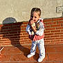 braided_hair, brick_wall, casual_clothing, child, daylight, doll, girl, happy, outdoor, pink_vest, plaster_wall, playful, portrait, shadow, smiling, standing, sunlight, toy, white_pants, white_shoes
