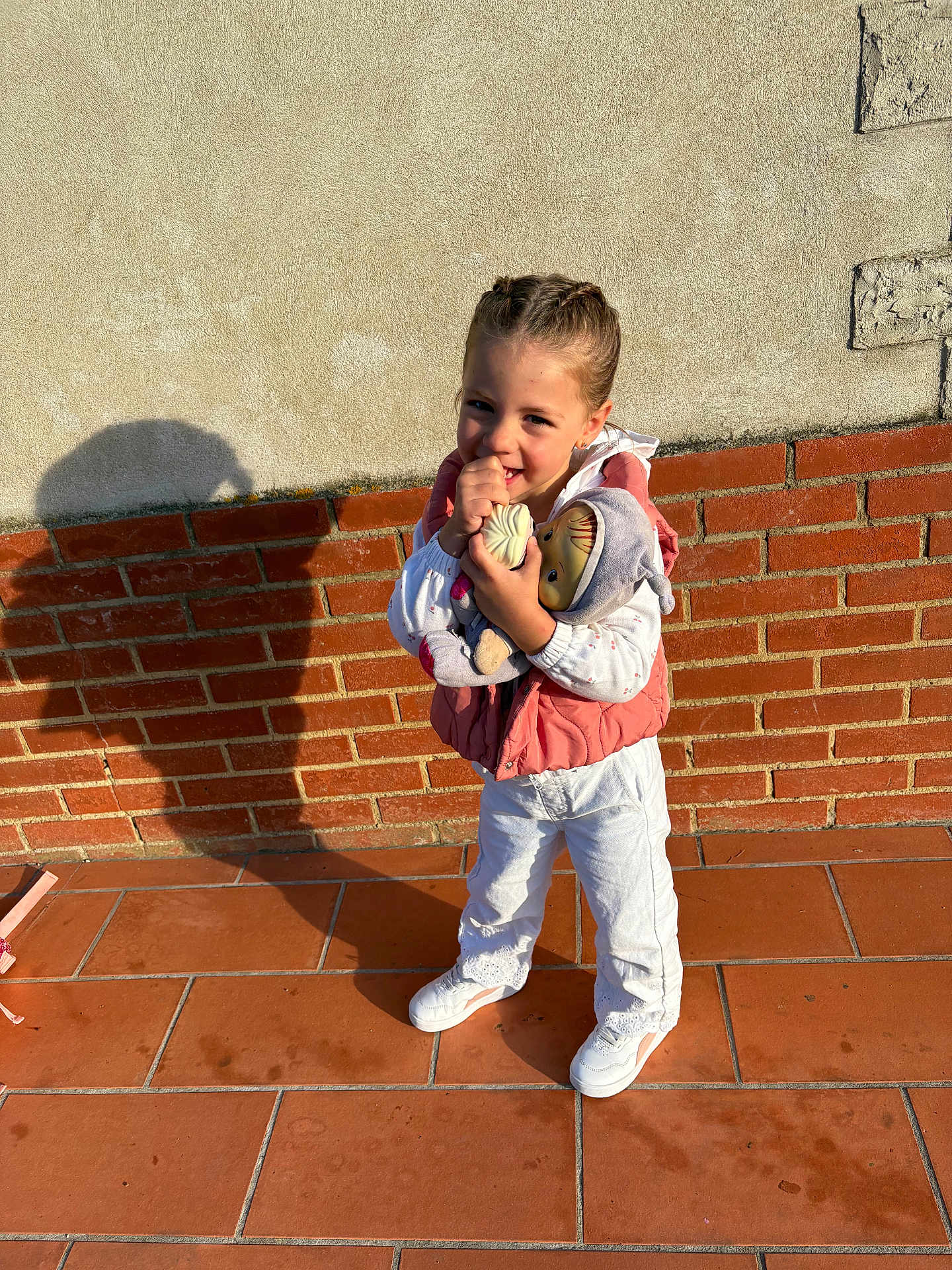 Maëlane a rejoint le concours — aidez-le/la à gagner de superbes lots ! braided_hair, brick_wall, casual_clothing, child, daylight, doll, girl, happy, outdoor, pink_vest, plaster_wall, playful, portrait, shadow, smiling, standing, sunlight, toy, white_pants, white_shoes