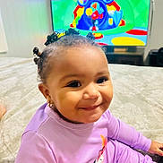 Aysha joined the competition — help win amazing prizes! toddler, child, smiling, indoor, bed, purple_clothing, cartoon, television, hair_buns, earrings, happy, cute, person, young_child, home, entertainment, playful, relaxed, portrait, cozy