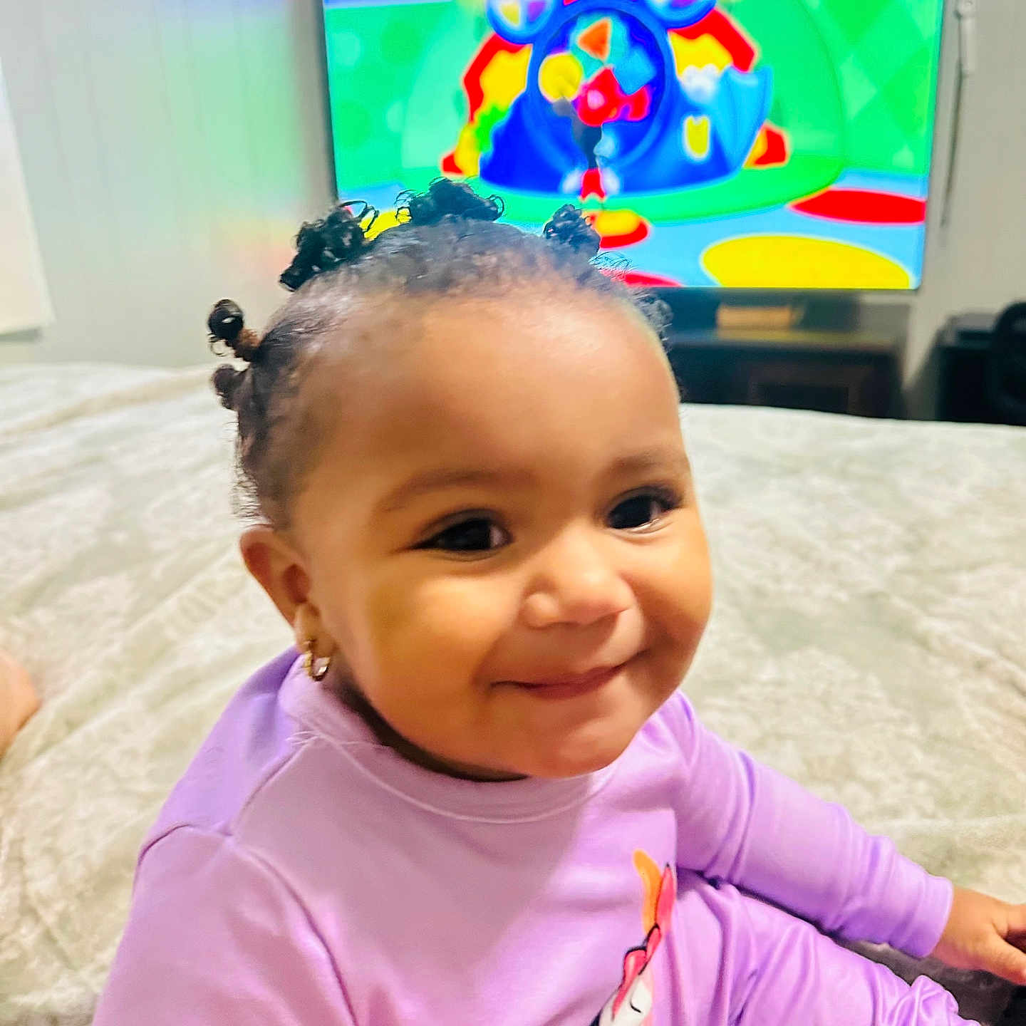 Aysha joined the competition — help win amazing prizes! bed, cartoon, child, cozy, cute, earrings, entertainment, hair_buns, happy, home, indoor, person, playful, portrait, purple_clothing, relaxed, smiling, television, toddler, young_child