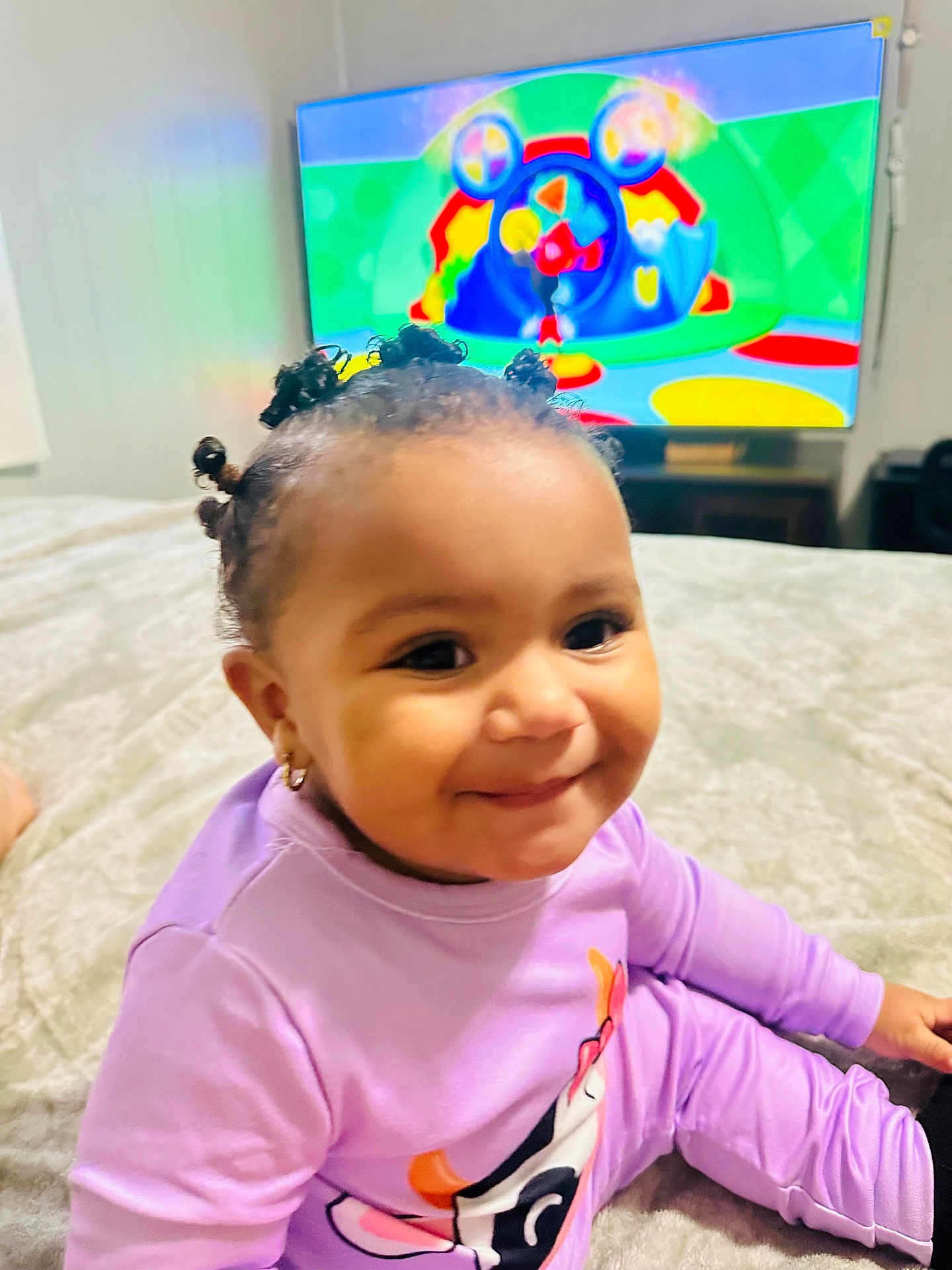 Aysha joined the competition — help win amazing prizes! toddler, child, smiling, indoor, bed, purple_clothing, cartoon, television, hair_buns, earrings, happy, cute, person, young_child, home, entertainment, playful, relaxed, portrait, cozy