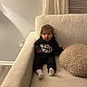 armchair, baby, black_clothing, blinds, brown_hair, child, couch, curtains, face, fluffy_pillow, home, indoors, pillow, portrait, sitting, sweatshirt, toddler, warm_lighting, white_socks, window