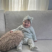 Elyanna is registered to the contest to win money with this photo: baby, bow, child, clothing, couch, curtains, cushion, happy, headband, home, indoor, infant, living_room, natural_light, patterned_pants, portrait, sitting, smiling, sofa, window