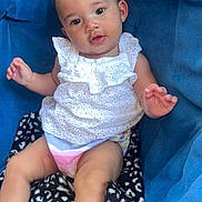 Leilani is registered to the contest to win money with this photo: baby, child, infant, sitting, portrait, smiling, face, eyes, white_outfit, dress, blue_cushion, blanket, indoor, seated, hand, legs, hair_bow, smartphone_screenshot, social_media_interface, casual_photo