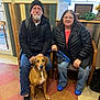 man, woman, dog, indoor, seated, glasses, black_jacket, blue_jeans, brown_dog, beanie, chairs, window, plants, wood_paneling, floor_tiles, leash, smiling, casual_clothing, pet, waiting_room