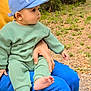 baby, barefoot, blue_cap, casual_clothing, child, curious, cute, daylight, grass, green_outfit, hand, infant, leaves, nature, outdoor, pants, person, side_view, sitting, young_child