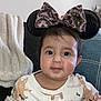 baby, child, headband, mickey_ears, bow, face, couch, blanket, indoor, cute, smile, long_sleeve, patterned_clothing, pink, white, cozy, person, portrait, young_child, happy