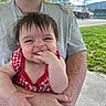 baby, child, person, smiling, happy, freckles, red_clothing, checkered_pattern, hand_in_mouth, outdoor, grass, sky, clouds, adult, holding, portrait, casual_clothing, daylight, nature, closeup
