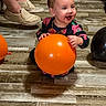 toddler, child, baby, smiling, happy, portrait, balloon, orange_balloon, black_balloon, indoor, wood_floor, sitting, playing, party, colorful_clothing, floral_clothing, necklace, adult_shoes, sneakers, photography