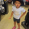 child, portrait, standing, hat, black_hat, white_shirt, shorts, pink_shoes, pouting, playful, halloween, costume_shop, skull, skeleton, retail_store, clothing, fashion, indoor, full_body, shopping