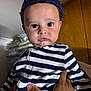 baby, child, striped_clothing, navy_cap, indoor, portrait, hand, curious_expression, onesie, face, person, young_child, holding, casual, looking_at_camera, pout, headwear, human, closeup, soft_lighting