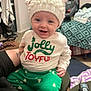 baby, blanket, car_seat, child, clothing, couch, furniture, green_pants, hand, happy, hat, indoor, jolly_and_joyful_shirt, knit_hat, person, portrait, remote_control, smiling, tattoo, white_hat