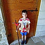 child, boy, wooden_door, cross, vest, red_ribbons, shorts, sneakers, stone_wall, tile_floor, indoors, smiling, standing, casual_clothing, playful, holding_object, young, portrait, background, light