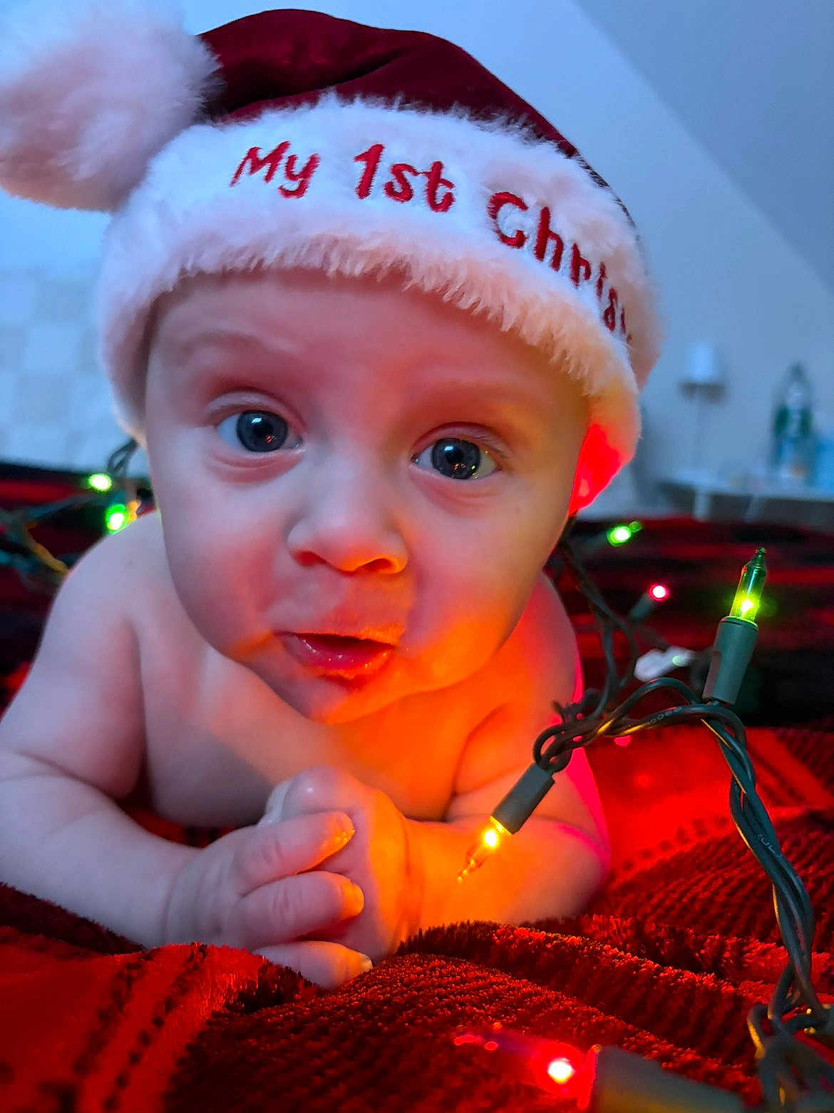 Luca is registered to the contest to win money with this photo: baby, infant, christmas_hat, holiday_lights, red_blanket, close_up, face, eyes, hands, indoors, festive, newborn, cute, expression, warm_lighting, celebration, child, portrait, holiday, seasonal