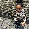 black_leggings, brick_wall, casual_clothing, child, concrete, curious, curly_hair, daytime, girl, hair_puff, hoodie, jacket, leopard_print, outdoor, person, sidewalk, sneakers, standing, urban, young