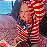 baby, child, hoodie, striped_clothing, blue_eyes, indoor, carpet, wicker_chair, ocean_view, beads, knitted_sweater, cute, sitting, curious, patterned_rug, furniture, toy, colorful, portrait, young_child