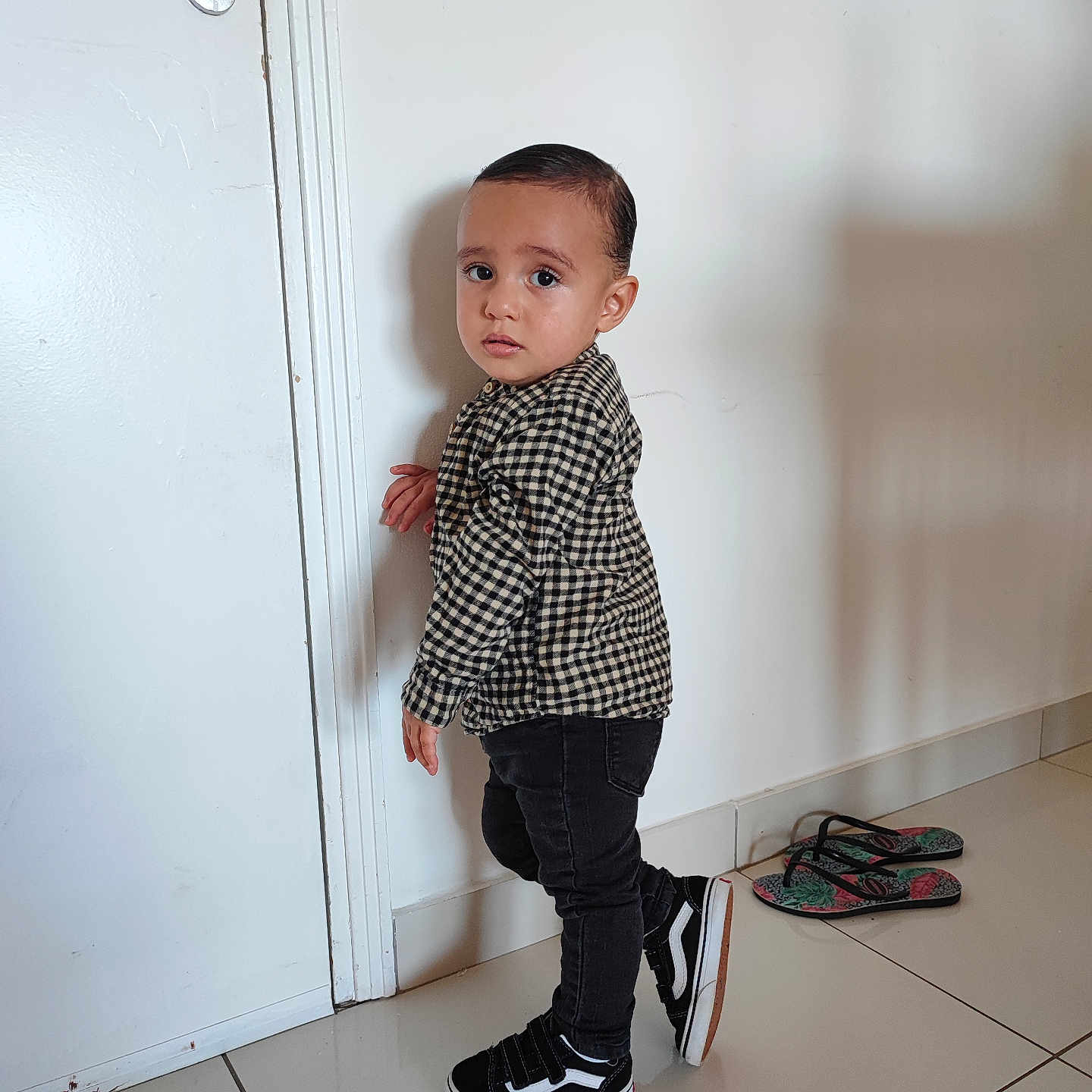Teïva a rejoint le concours — aidez-le/la à gagner de superbes lots ! black_jeans, black_shoes, checkered_shirt, child, clothing, curious_expression, door, flip_flops, floor, footwear, hair, home, indoor, person, shadow, standing, tile_floor, toddler, white_wall, young_child