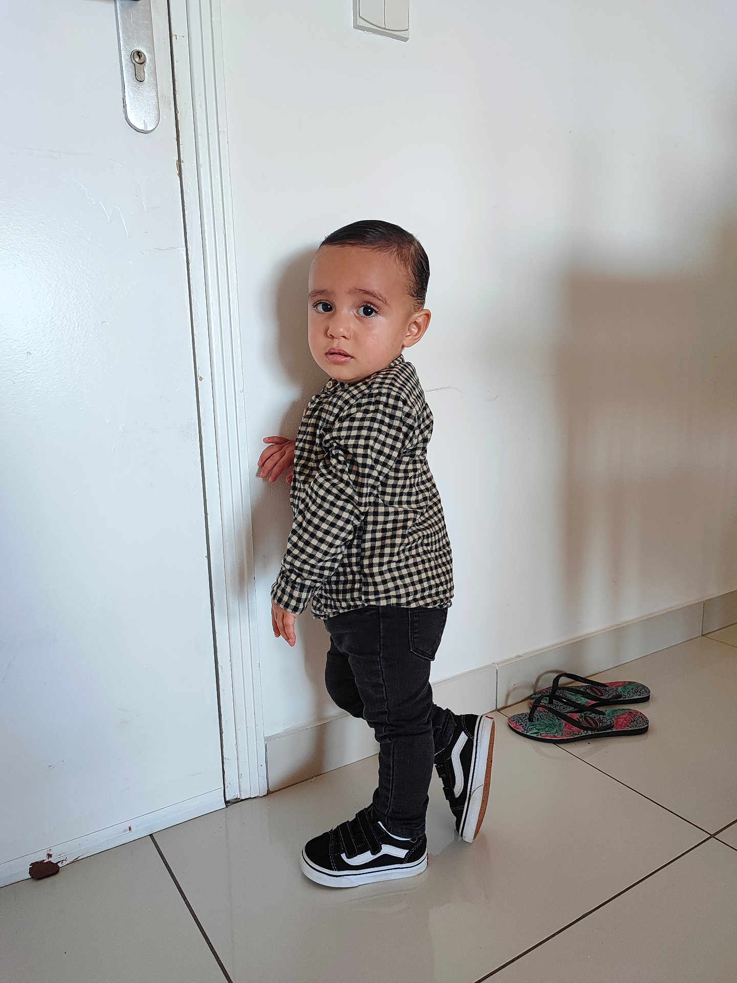 Teïva a rejoint le concours — aidez-le/la à gagner de superbes lots ! toddler, child, indoor, standing, checkered_shirt, black_jeans, black_shoes, door, white_wall, tile_floor, flip_flops, footwear, curious_expression, person, young_child, hair, clothing, shadow, floor, home