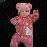 Meadow is registered to the contest to win money with this photo: baby, infant, child, portrait, lying_down, pink_outfit, onesie, polka_dots, animal_hat, bear_hat, sheep_design, toy, blanket, black_background, indoors, waving, sitting, facial_expression, full_body, cozy