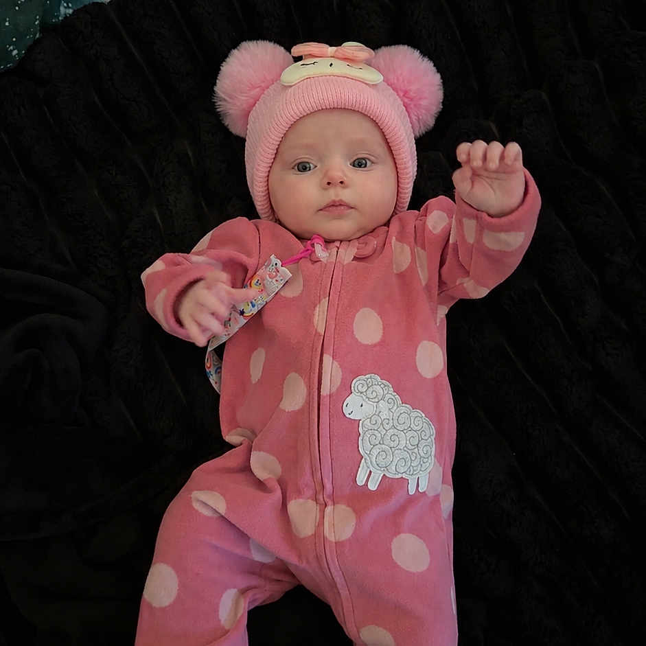 Meadow is registered to the contest to win money with this photo: baby, infant, child, portrait, lying_down, pink_outfit, onesie, polka_dots, animal_hat, bear_hat, sheep_design, toy, blanket, black_background, indoors, waving, sitting, facial_expression, full_body, cozy