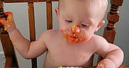 Liam joined the competition — help win amazing prizes! baby, birthday_cake, cake, celebration, child, face, food, frosting, hands, happy, high_chair, indoor, messy, person, plate, red_frosting, smudged, table, wooden_chair, yellow_frosting