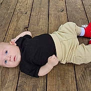 Liam is registered to the contest to win money with this photo: baby, infant, child, wooden_deck, floor, red_shoes, black_shirt, beige_pants, lying_down, outdoor, curious, blue_eyes, casual_clothing, portrait, person, cute, small_hands, relaxed, daylight, skin