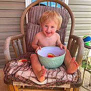 Liam is registered to the contest to win money with this photo: toddler, child, rocking_chair, watermelon, bowl, porch, window, blue_sky, ball, cushion, wooden_chair, smiling, barefoot, messy_face, summer, outdoor, happy, person, food, casual