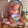 toddler, child, rocking_chair, watermelon, bowl, porch, window, blue_sky, ball, cushion, wooden_chair, smiling, barefoot, messy_face, summer, outdoor, happy, person, food, casual